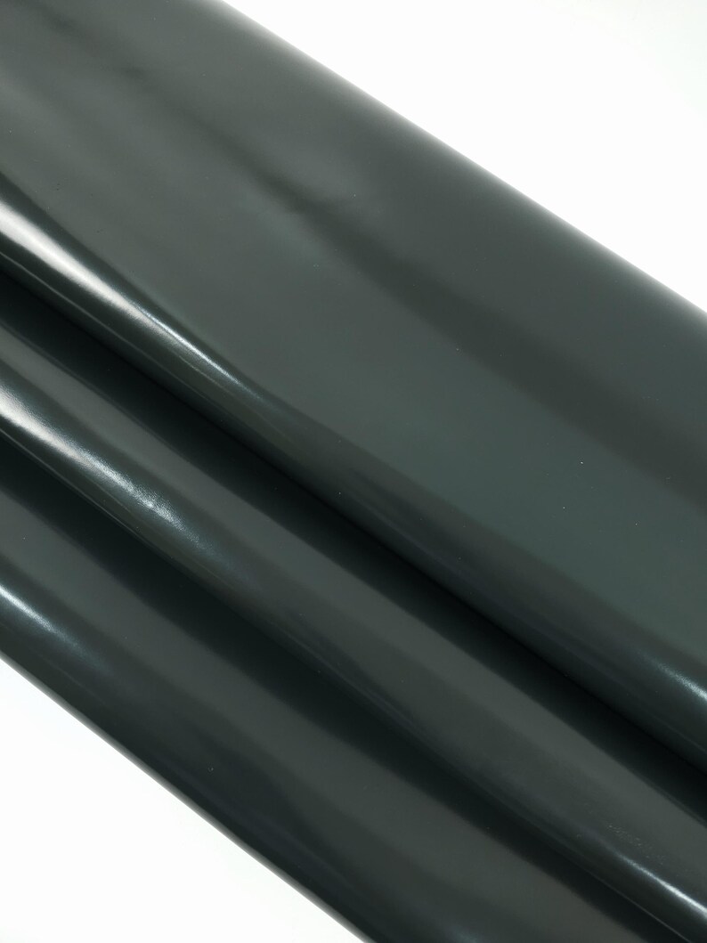 May include: A roll of dark gray vinyl fabric with a glossy finish. The material is folded over itself, creating multiple layers and highlighting the sheen of the surface. The fabric appears smooth and reflective, suitable for various crafting projects.