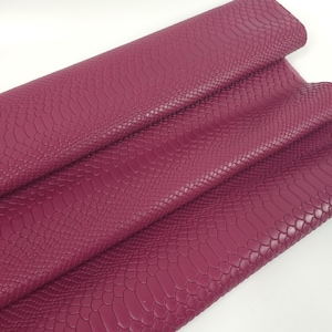 Può includere: Un rotolo di tessuto in similpelle color magenta caratterizzato da una texture in rilievo che imita la pelle di serpente. Il materiale è ripiegato su se stesso, mostrando un motivo a scaglie uniforme sulla superficie del foglio flessibile.