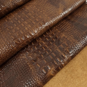 Puede incluir: Primer plano de cuero marrón con textura de cocodrilo. El cuero tiene un patrón detallado con una superficie rugosa y texturizada. El color es un marrón oscuro intenso con reflejos marrones más claros, lo que sugiere un acabado natural. La imagen muestra varias capas de cuero.