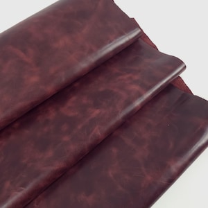 May include: A folded piece of deep burgundy leather with a smooth, matte finish and subtle, darker marbled color variations across the surface. The material shows a soft, pliable texture with visible grain patterns and a rich, dark red tone.