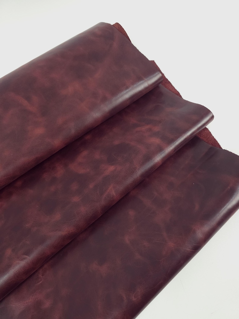 May include: A folded piece of deep burgundy leather with a smooth, matte finish and subtle, darker marbled color variations across the surface. The material shows a soft, pliable texture with visible grain patterns and a rich, dark red tone.