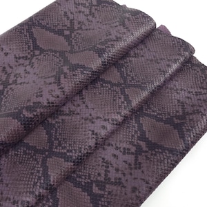 May include: Close-up of a folded piece of purple snakeskin leather. The leather has a scale pattern in shades of purple and black. The material is likely used for crafting items like bags or accessories.