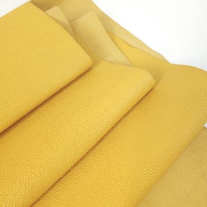 May include: Close-up of several sheets of yellow leather. The leather has a textured, bumpy surface. The sheets are layered, revealing different shades of yellow, from a bright, sunny hue to a softer, paler tone. The leather appears to be new and unused.