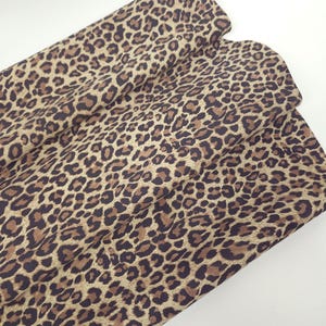 Könnte beinhalten: Ein gefaltetes Stück Stoff mit einem sich wiederholenden Leopardenmuster. Das Design besteht aus dunkelbraunen und schwarzen Flecken auf einem hellbraunen Hintergrund, was eine klassische Tierfelloptik auf der Textiloberfläche erzeugt.
