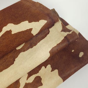 May include: A folded piece of cowhide with a natural pattern. The hide features a mix of brown and cream colors, with the brown forming irregular shapes against the cream background. The texture appears soft and the overall look is rustic.