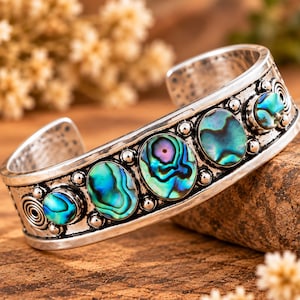 Puede incluir: Un brazalete de plata tipo puño con cinco incrustaciones ovaladas de concha de abulón en tonos azules y verdes iridiscentes. La banda de metal está decorada con pequeñas esferas de plata y patrones en espiral alrededor de cada incrustación.