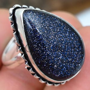 May include: A silver ring featuring a large, teardrop-shaped, dark blue gemstone with a speckled, glittery effect. The ring has a silver band and a decorative silver border around the gemstone. The text "MG JEWELRY" is visible in the top left corner.