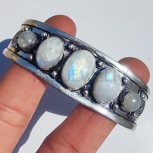 May include: A silver cuff bracelet with five oval moonstone cabochons. The bracelet has a detailed design with small silver beads and spiral accents. The text "MG JEWELRY" is visible in the upper left corner.