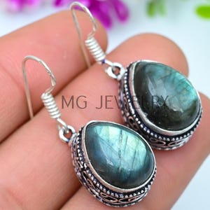 Puede incluir: Pendientes plateados con piedras de labradorita iridiscentes en forma de lágrima. Las piedras están engastadas en biseles detallados y ornamentados. Cierre de gancho. El texto "MG JEWELRY" es visible.