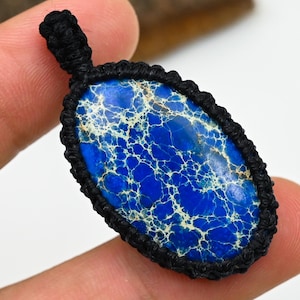 May include: A blue and white stone pendant with a black woven border and loop. The stone has a unique pattern of blue and white veins. The pendant is held by a finger.