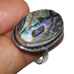 Puede incluir: Un anillo de plata con una incrustación ovalada de concha de abulón. La concha tiene colores iridiscentes de verde, azul, morado y marrón.