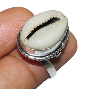 Puede incluir: Un anillo de plata con una concha de cauri blanca en el centro. El anillo tiene un diseño de cuentas alrededor de la concha.