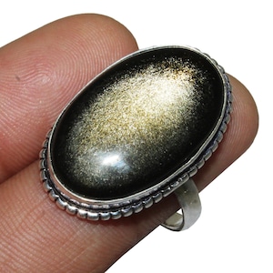 Peut inclure: Une bague en argent avec une grande pierre précieuse ovale noire avec des reflets dorés. La bague a un bord perlé.