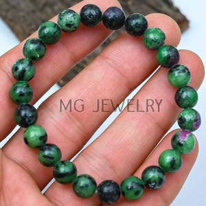 May include: A green and black gemstone bracelet with a pink accent. The bracelet is made of round beads and is strung on a clear elastic cord.
