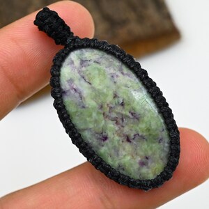 Può includere: Ciondolo ovale con una pietra verde e viola, incastonata in una cornice di macramè nero. Il ciondolo ha un anello nella parte superiore per essere appeso. La pietra ha una superficie naturale e strutturata.