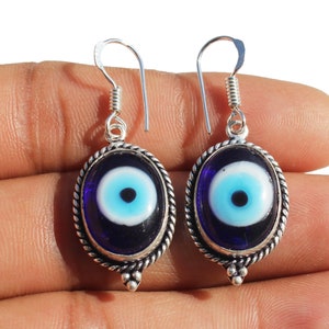 May include: A pair of silver earrings with blue evil eye charms. The charms are oval shaped and have a white center with a black pupil. The earrings are hanging from silver hooks.