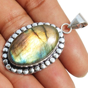 May include: A silver pendant with a large oval-shaped labradorite gemstone. The gemstone has a iridescent sheen with shades of green, blue, and gold. The pendant is surrounded by a silver beaded border.