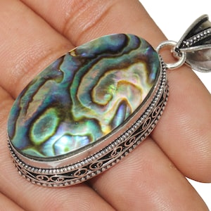 Könnte beinhalten: Ein silberner Anhänger mit einem ovalen Einlegeteil aus Abalone-Muschel. Die Muschel hat schillernde grüne, blaue und violette Farben. Der Anhänger hat einen detaillierten silbernen Rand.