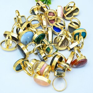 Puede incluir: Una colección de anillos dorados con varias piedras preciosas ovaladas de colores. Los anillos están dispuestos en una pila sobre una superficie blanca.