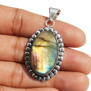 May include: Silver pendant featuring a large oval-shaped labradorite gemstone with iridescent green, gold, and blue hues. The stone is set in a silver bezel with a beaded border.