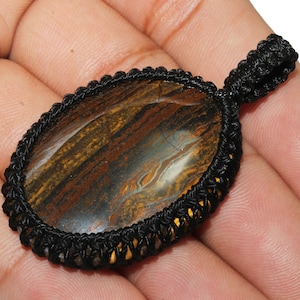 Może przedstawiać: Owalny wisiorek z brązowego i złotego kamienia szlachetnego owinięty czarną sznurówką. Wisiorek jest osadzony w czarnej sznurówce z węzłowym wzorem.