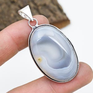 May include: Oval-shaped pendant with a polished, banded agate stone in shades of gray and white. The stone is set in a silver-colored metal frame with a loop for a chain. The pendant is approximately 1.5 inches long.