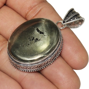 Puede incluir: Colgante ovalado de pirita con un engaste de metal plateado. La pirita tiene un brillo metálico y una superficie texturizada. El engaste presenta un diseño ornamentado en los bordes y una argolla decorativa.
