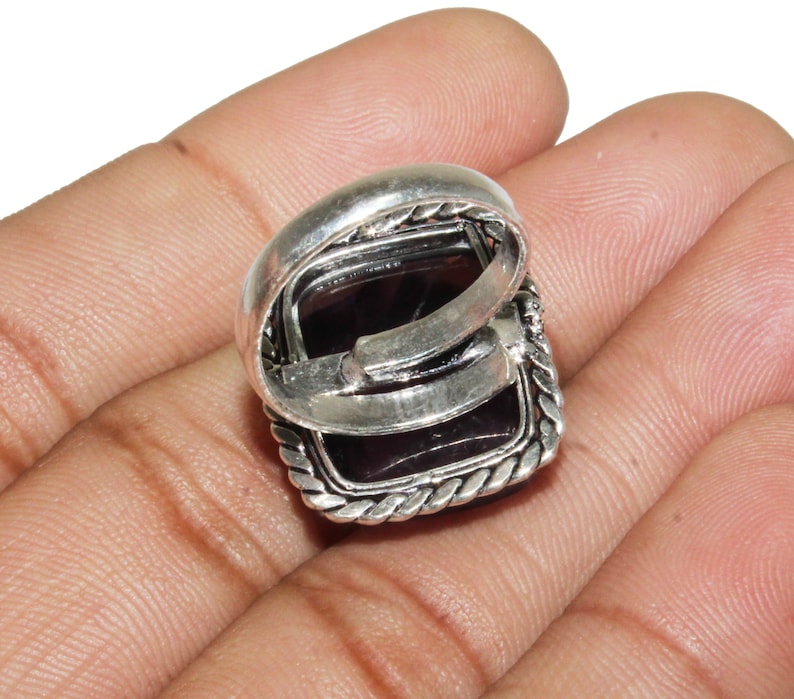May include: A silver ring with a square-shaped purple gemstone set in a silver bezel. The ring has a decorative rope-like border around the gemstone.