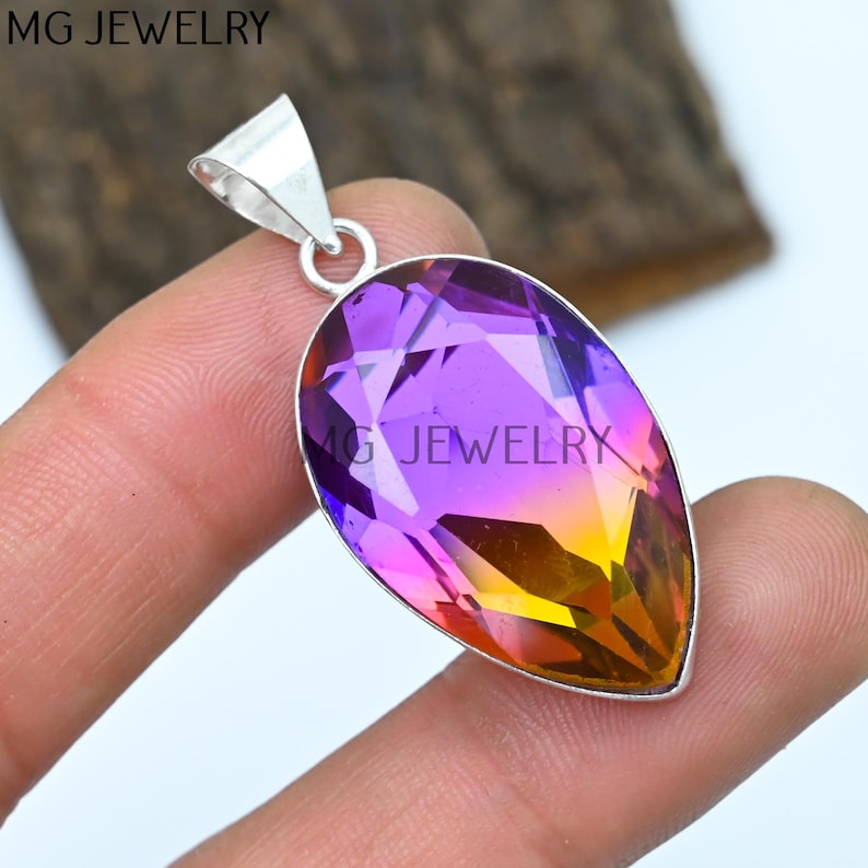 May include: A teardrop-shaped pendant with a purple, pink, and orange gemstone set in sterling silver. The pendant is on a finger.