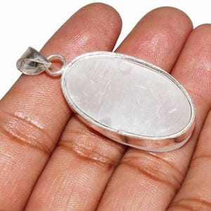 May include: A silver oval pendant with a white selenite stone. The pendant is on a hand.