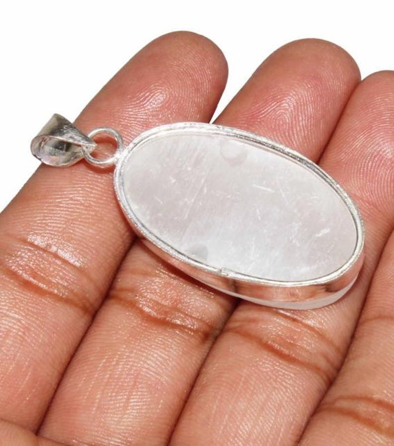 May include: A silver oval pendant with a white selenite stone. The pendant is on a hand.