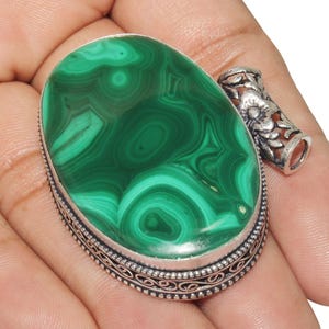 Puede incluir: Un colgante de piedra de malaquita verde con un diseño de filigrana de plata. El colgante tiene forma ovalada y una superficie lisa y pulida.