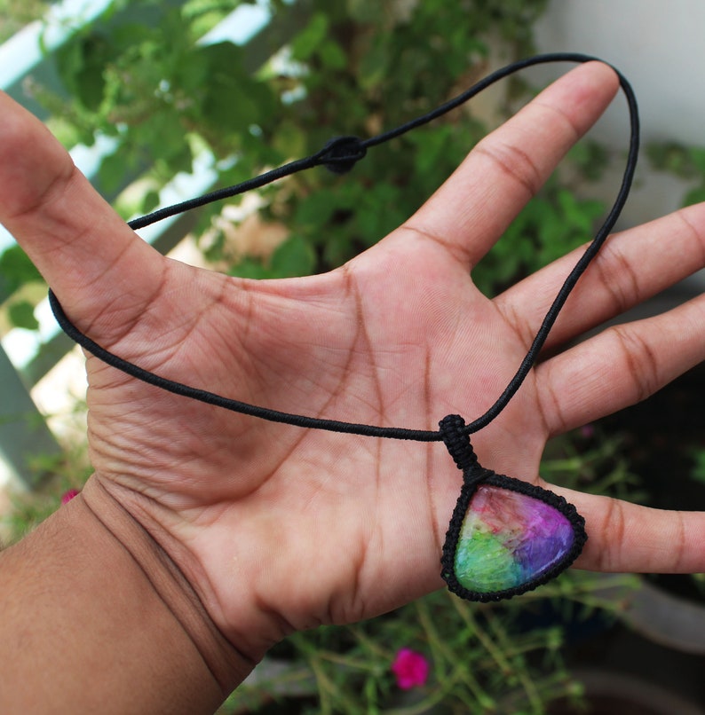 May include: A rainbow-colored gemstone pendant on a black cord necklace. The pendant is a teardrop shape and has a variety of colors, including pink, purple, green, and blue.