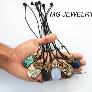 May include: Six different pendants with various natural stone inlays, each with a carved design and a black cord. The pendants are held in a hand. The text "MG JEWELRY" appears in the image.