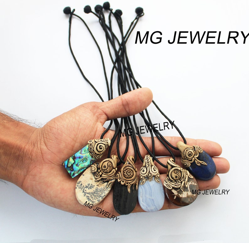 May include: Six different pendants with various natural stone inlays, each with a carved design and a black cord. The pendants are held in a hand. The text "MG JEWELRY" appears in the image.