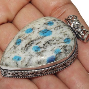 Puede incluir: Un colgante de plata con una piedra preciosa de cianita blanca y azul. El colgante tiene un diseño de plata intrincado alrededor del borde.