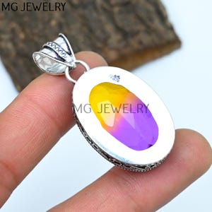 May include: A silver pendant with an oval-shaped, yellow and purple gemstone. The pendant has an ornate silver bail and a detailed silver border. The back of the pendant is engraved with the text "MG JEWELRY".