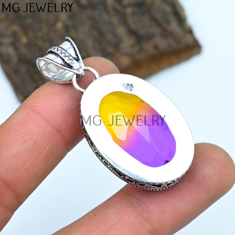 May include: A silver pendant with an oval-shaped, yellow and purple gemstone. The pendant has an ornate silver bail and a detailed silver border. The back of the pendant is engraved with the text "MG JEWELRY".