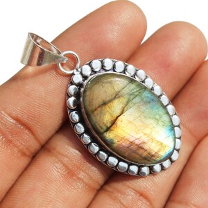 May include: A silver pendant with a large oval-shaped labradorite gemstone. The gemstone has a iridescent sheen with shades of green, blue, and gold. The pendant is surrounded by a silver beaded border.