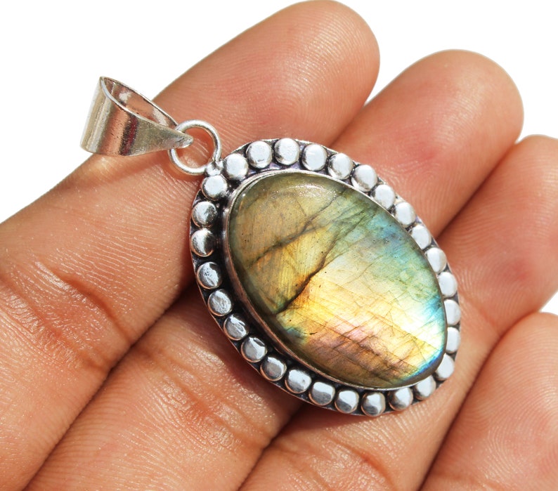 May include: A silver pendant with a large oval-shaped labradorite gemstone. The gemstone has a iridescent sheen with shades of green, blue, and gold. The pendant is surrounded by a silver beaded border.