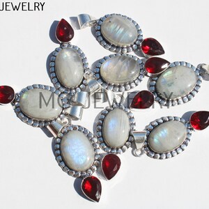 May include: A group of nine silver pendants with oval-shaped moonstone gemstones and red garnet accents.  MG JEWELRY is printed in the top left corner of the image.