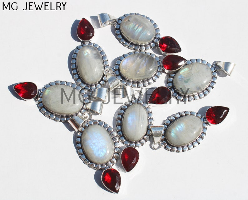 May include: A group of nine silver pendants with oval-shaped moonstone gemstones and red garnet accents.  MG JEWELRY is printed in the top left corner of the image.