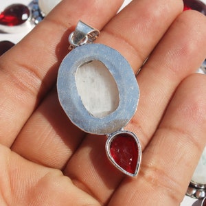 May include: Silver pendant with a white oval gemstone and a red teardrop gemstone. The pendant is on a hand.