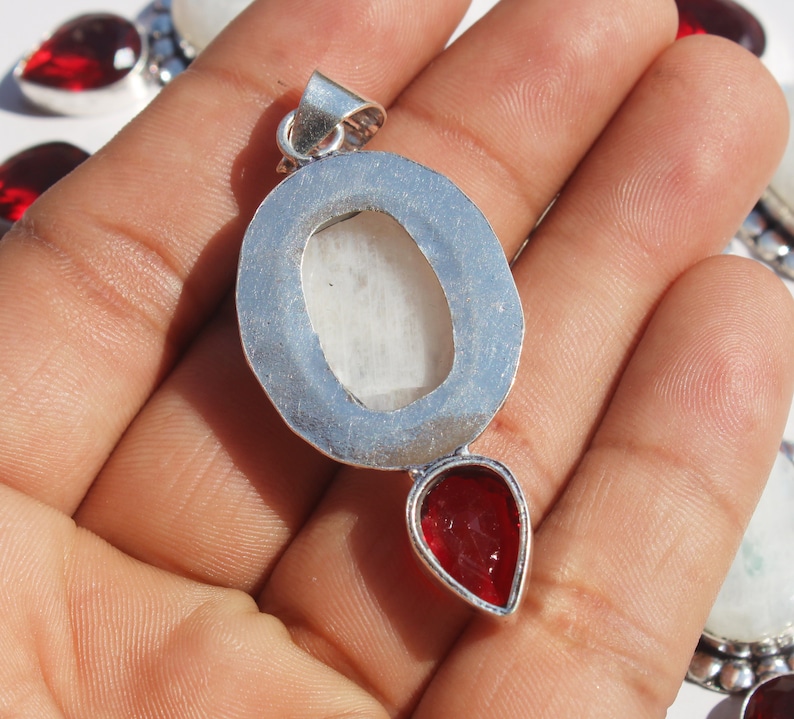May include: Silver pendant with a white oval gemstone and a red teardrop gemstone. The pendant is on a hand.