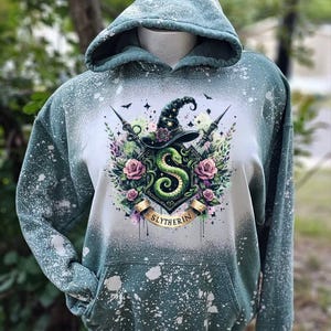 May include: A green hooded sweatshirt with a white bleach splattered design. The design features a green snake, a witch's hat, and the words "Slytherin".
