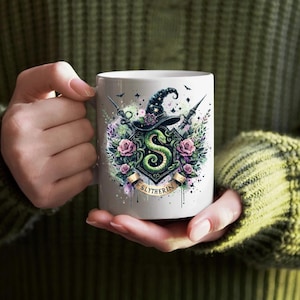 May include: White ceramic mug with a green snake and a black hat design. The design includes the words "Slytherin" and floral accents.