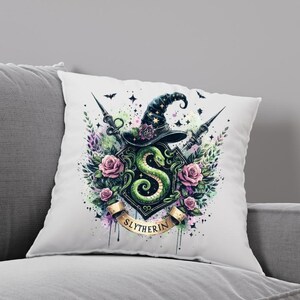 May include: A white pillow with a watercolor illustration of the Slytherin house crest from the Harry Potter series. The crest features a green snake, a wizard's hat, and the words "Slytherin" in gold lettering.