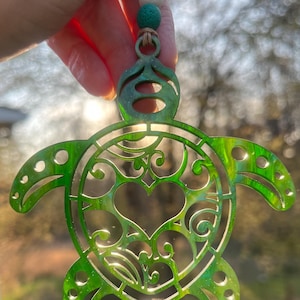 May include: A green, decorative turtle ornament with a heart-shaped design in the center. The ornament is made of a translucent material, with a beaded string for hanging. The turtle's shell features intricate cut-out patterns.