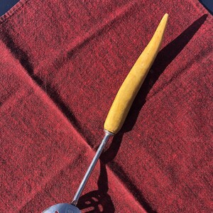 May include: A vintage kitchen slotted spoon with a yellow wooden handle and a stainless steel head. The spoon is resting on a red textured cloth, showcasing its design and materials. The spoon's head has a series of cut-out slots.