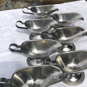 May include: A collection of six silver-toned stainless steel gravy boats arranged in a staggered formation. Each boat features a polished finish, a curved handle, a wide pouring spout, and a circular pedestal base with a decorative beaded edge.